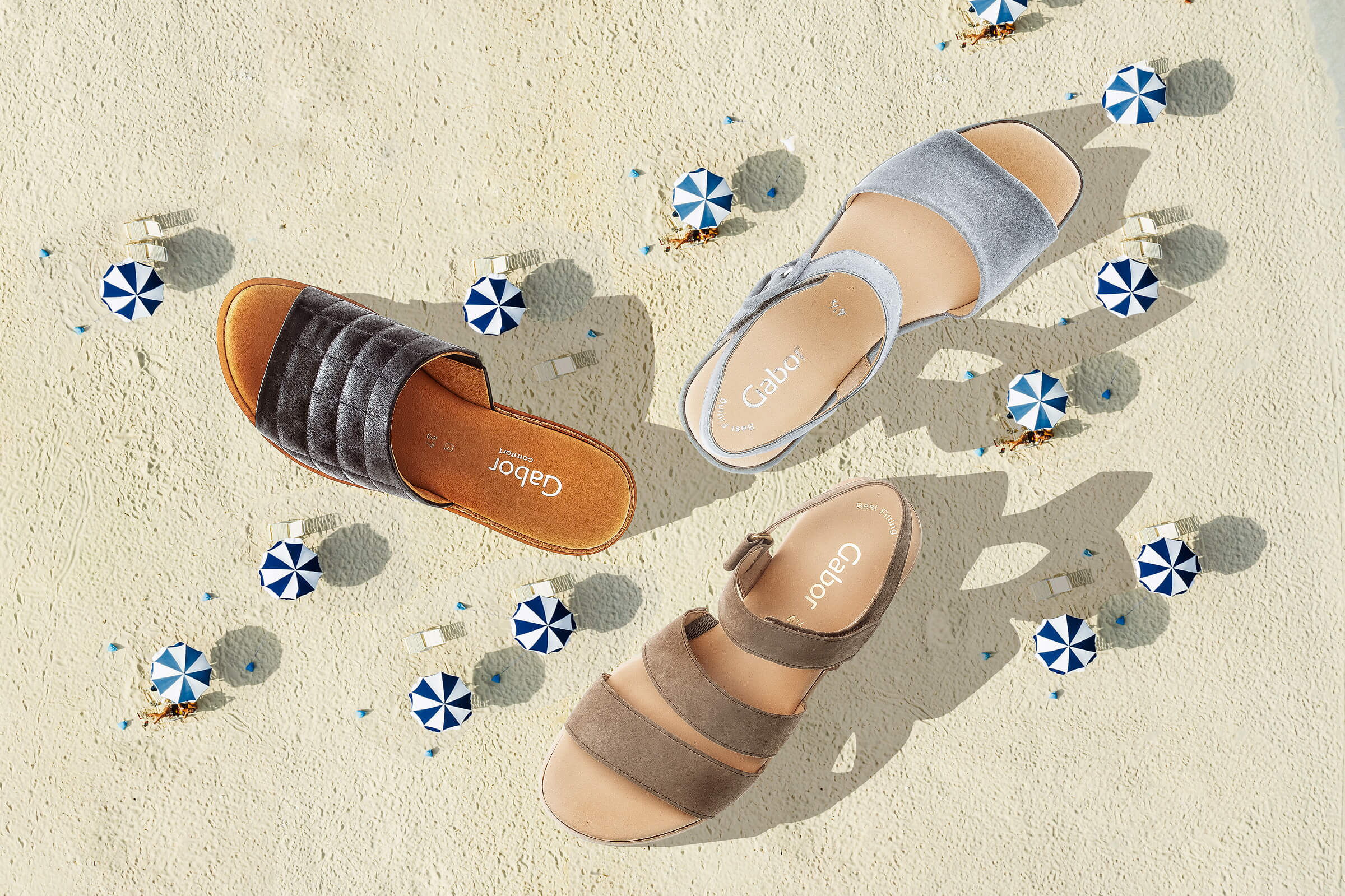 Werbefotografie mit Sandstrand mit Sonnenschirmen und drei Sommersandalen von oben